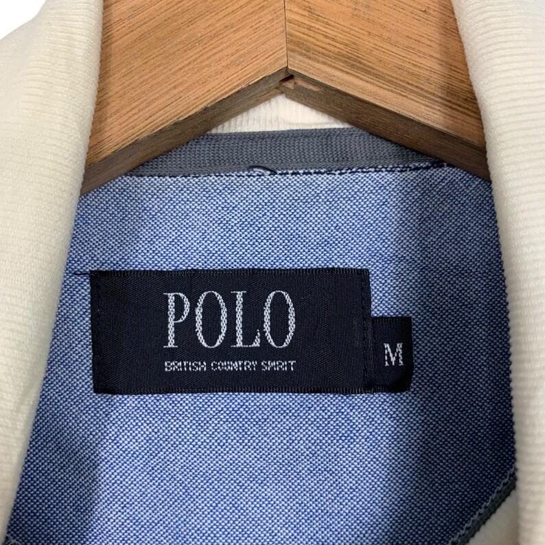 May include: Close-up of a blue button-down shirt with a white corduroy collar. The shirt features a black label with the word "POLO" and the text "BRITISH COUNTRY SPIRIT". The size "M" is also visible on the label.