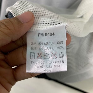 Puede incluir: Una etiqueta blanca con texto negro que dice "FM 6404", "表地 ポリエステル 100%", "裏地 ポリエステル 100%", "ダイワボウアドバンス株式会社", "TEL 03-4332-5231" y "中国製".