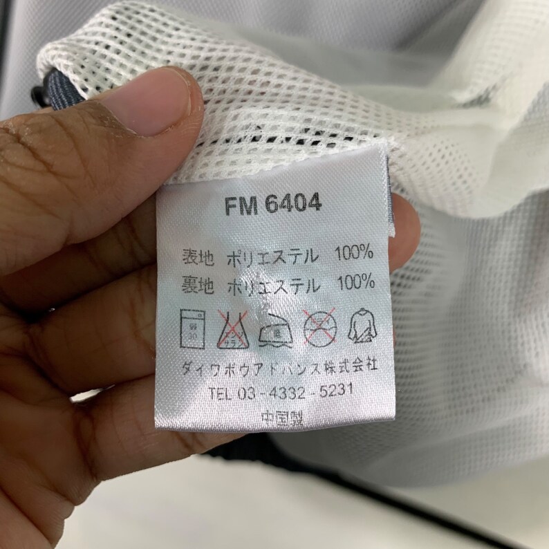 Puede incluir: Una etiqueta blanca con texto negro que dice "FM 6404", "表地 ポリエステル 100%", "裏地 ポリエステル 100%", "ダイワボウアドバンス株式会社", "TEL 03-4332-5231" y "中国製".