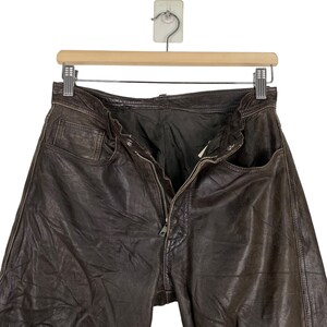 May include: Brown leather pants with a zipper fly and belt loops. The pants are hanging on a wooden hanger.