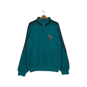 May include: Teal blue fleece pullover sweatshirt with a colorful cartoon character embroidered on the front. The sweatshirt has a mock neck and a single button closure. The sleeves have a geometric pattern in black, purple, and white.