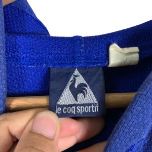 Puede incluir: Una sudadera con capucha azul con una etiqueta negra y plateada que dice "le coq sportif" y tiene un logotipo de gallo.