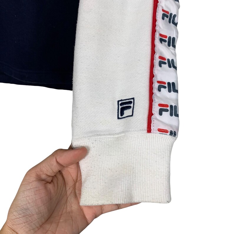 May include: Close-up of a white and navy blue sweatshirt sleeve. The white sleeve features a navy blue logo and a red and white stripe with the brand name. The cuff is white.