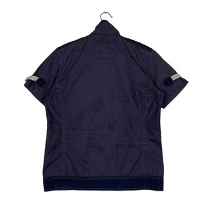 May include: A navy blue short-sleeved shirt with a zip fastening and a contrasting navy blue trim around the collar and cuffs. The shirt has two pockets on the front and a patch on each sleeve.