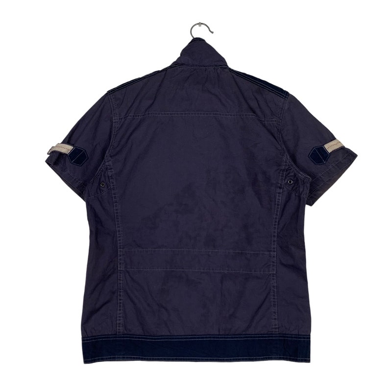 May include: A navy blue short-sleeved shirt with a zip fastening and a contrasting navy blue trim around the collar and cuffs. The shirt has two pockets on the front and a patch on each sleeve.