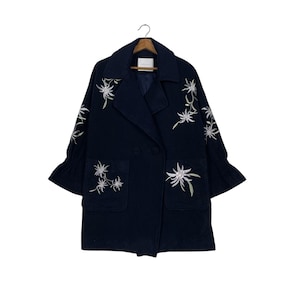 May include: A navy blue coat with white floral embroidery. The coat has a double-breasted design, two front pockets, and flared sleeves. The coat is hanging on a wooden hanger against a white backdrop.