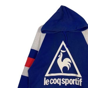 Puede incluir: Una sudadera con capucha azul y gris con una franja roja y blanca en la manga. La sudadera presenta un logotipo de gallo blanco en un tri&aacute;ngulo con el texto "le coq sportif" debajo.