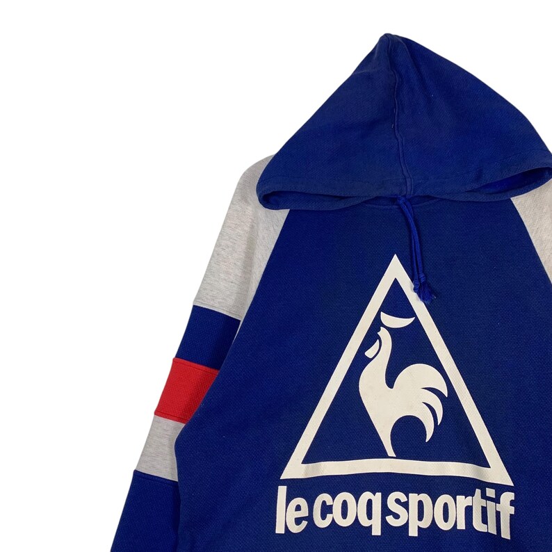 Puede incluir: Una sudadera con capucha azul y gris con una franja roja y blanca en la manga. La sudadera presenta un logotipo de gallo blanco en un tri&aacute;ngulo con el texto "le coq sportif" debajo.