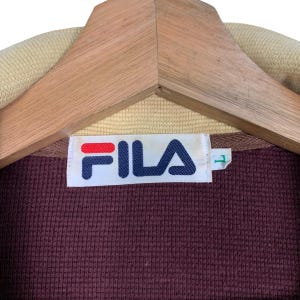 May include: Close-up of a maroon FILA jacket label. The label is white with the FILA logo in red and blue. The jacket is hanging on a wooden hanger. The collar is tan.