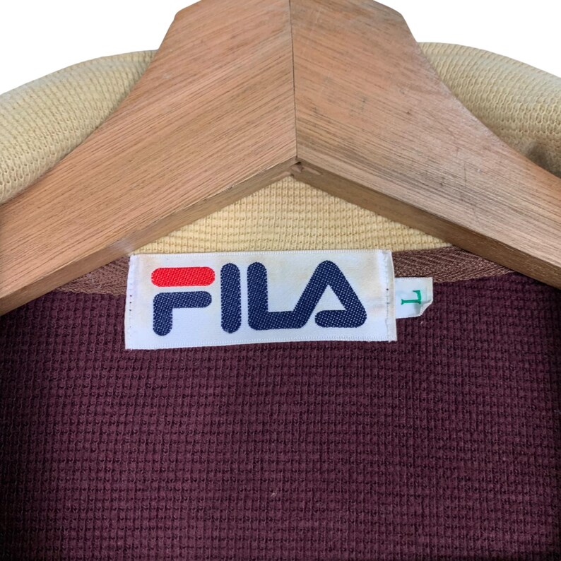 May include: Close-up of a maroon FILA jacket label. The label is white with the FILA logo in red and blue. The jacket is hanging on a wooden hanger. The collar is tan.
