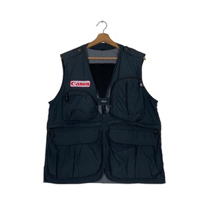 Op de afbeelding: Een donkerblauw vest met meerdere zakken en een Canon-logo op de voorkant. Het vest heeft een mesh-rugpaneel en een ritssluiting.