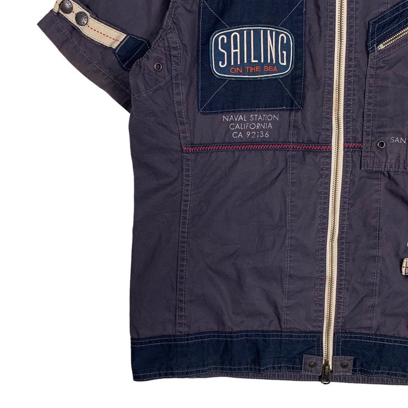 May include: A dark blue short-sleeved jacket with a zip fastening. The jacket has a patch on the chest that says "Sailing on the Sea" and "Naval Station California CA 92136".