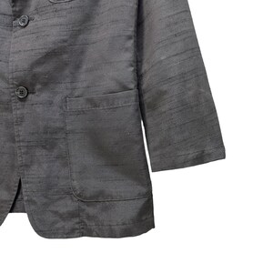 Puede incluir: Una chaqueta negra con tejido texturizado y dos botones de cierre. La chaqueta tiene un bolsillo de parche en el lado izquierdo y un bolsillo con solapa en el lado derecho.