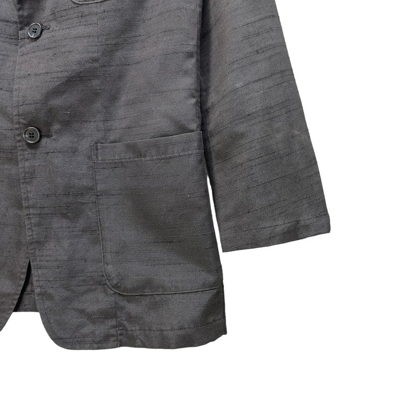 Puede incluir: Una chaqueta negra con tejido texturizado y dos botones de cierre. La chaqueta tiene un bolsillo de parche en el lado izquierdo y un bolsillo con solapa en el lado derecho.