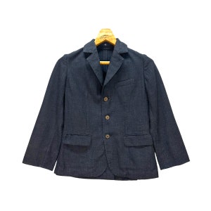 Könnte beinhalten: Ein dunkelblauer Blazer mit fallendem Revers, drei Knöpfen und zwei Vordertaschen. Der Blazer hängt an einem Holzkleiderbügel. Der Stoff hat eine subtile Textur.