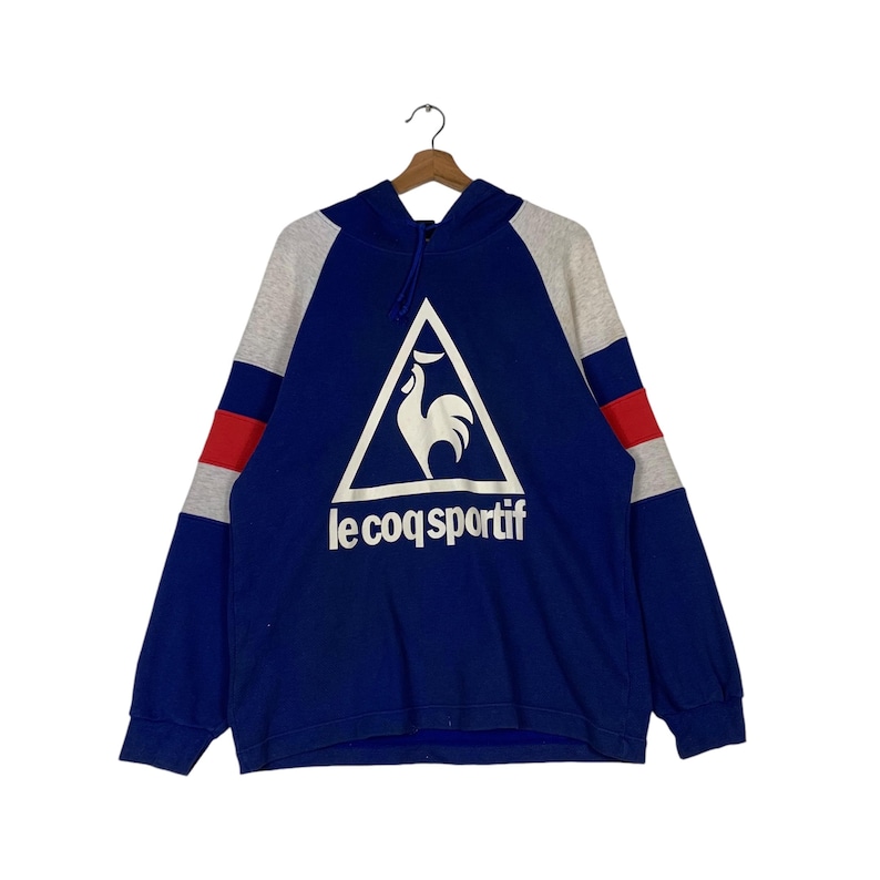 Puede incluir: Una sudadera con capucha azul con un logotipo de gallo blanco en un tri&aacute;ngulo y el texto "le coq sportif" en blanco. La sudadera tiene rayas grises y rojas en las mangas.