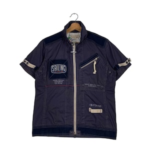 May include: A dark blue short-sleeved jacket with a zip fastening. The jacket has several patches with text, including "Sailing on the Sea", "Naval Station California CA 93106", "Santa Monica 1960", and "CR2 PTERAVES".