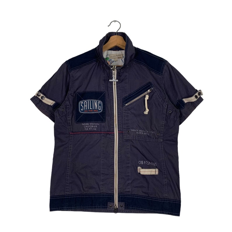 May include: A dark blue short-sleeved jacket with a zip fastening. The jacket has several patches with text, including "Sailing on the Sea", "Naval Station California CA 93106", "Santa Monica 1960", and "CR2 PTERAVES".