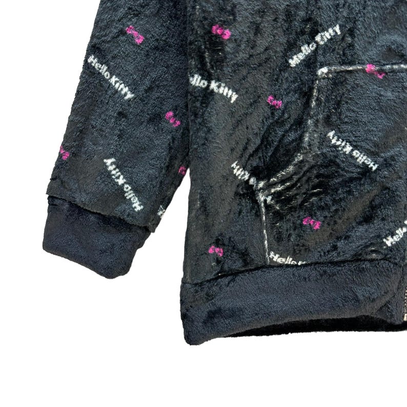 Może przedstawiać: Czarna pluszowa bluza z kapturem z białym napisem "Hello Kitty" i r&oacute;żowymi akcentami w postaci kokardek. Bluza z kapturem posiada przednią kieszeń i ciemny mankiet. Miękki, puszysty materiał jest widoczny na tym zbliżeniu.