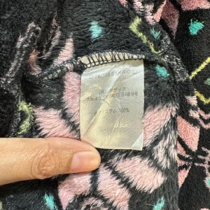 May include: Close-up of a black and pink fabric with a white tag. The tag has Japanese text and the logo "'76,'10 SANRIO." The fabric has a fuzzy texture.