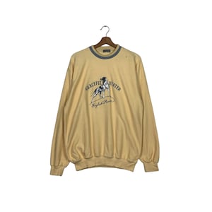 May include: A light yellow sweatshirt with a crew neck and long sleeves. The front features a graphic of a dog with the text "GRACEFUL HUNTER" and "English Pointer" in a black and white design. The collar is trimmed with a blue-gray color.