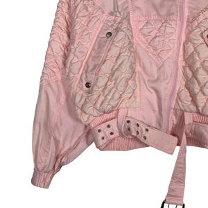 May include: A pink quilted jacket with a zip fastening and a belt. The jacket has a hood and a logo on the back.