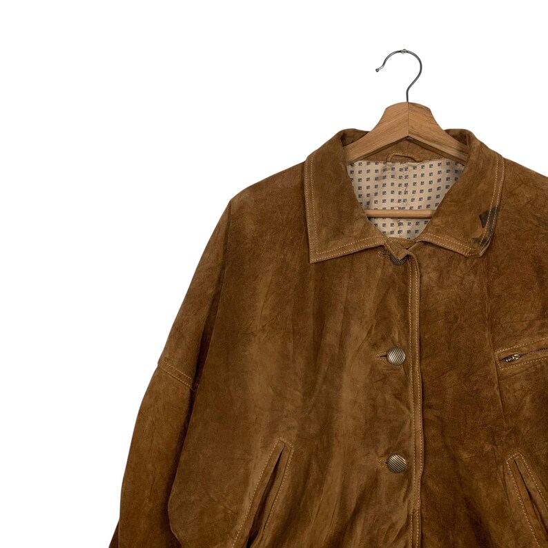 May include: A brown suede jacket with a button-up front, a collar, and two front pockets. The jacket is hanging on a wooden hanger against a white background. The jacket has a textured appearance.