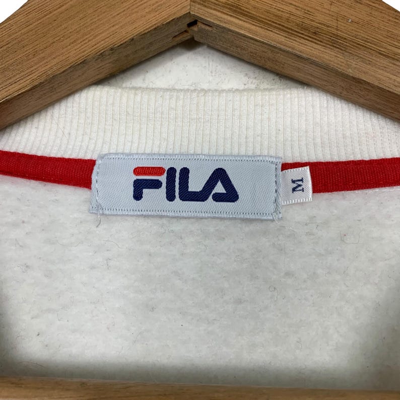 May include: Close-up of a white sweatshirt collar with a red stripe. The Fila logo is centered on a light blue tag. The tag also has the letter "M" on it. The sweatshirt is on a wooden hanger.