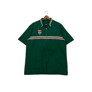 Puede incluir: Camisa verde de manga corta con cremallera completa. La camisa presenta un pequeño bolsillo en el pecho y un diseño de rayas horizontales en rojo, blanco y naranja. El logotipo de "7-Eleven" está en el pecho izquierdo.