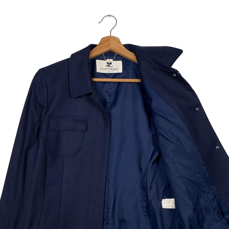 May include: A navy blue jacket with a collared neckline and a single pocket on the left side. The jacket is hanging on a wooden hanger. The interior lining is a darker shade of blue. The brand name "Courr&egrave;ges" is visible on the label.