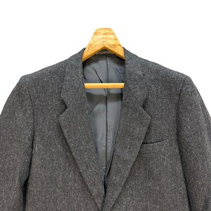 Puede incluir: Una chaqueta gris con un patr&oacute;n texturizado cuelga de una percha de madera. La chaqueta tiene un cierre de un bot&oacute;n y un bolsillo en el lado izquierdo.