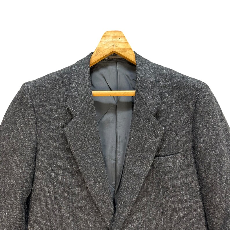 Puede incluir: Una chaqueta gris con un patr&oacute;n texturizado cuelga de una percha de madera. La chaqueta tiene un cierre de un bot&oacute;n y un bolsillo en el lado izquierdo.