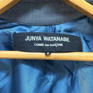 Puede incluir: Una etiqueta negra con texto blanco que dice "JUNYA WATANABE COMME des GARCONS" sobre un fondo de tela azul. La etiqueta est&aacute; unida a una prenda con una peque&ntilde;a etiqueta de talla que dice "S".