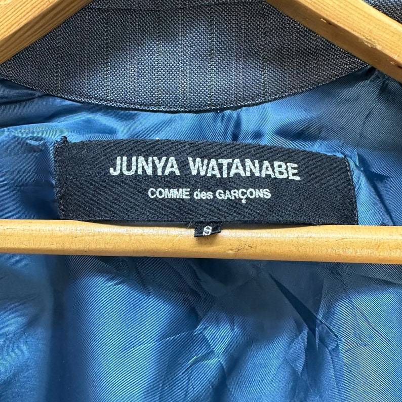 Puede incluir: Una etiqueta negra con texto blanco que dice "JUNYA WATANABE COMME des GARCONS" sobre un fondo de tela azul. La etiqueta est&aacute; unida a una prenda con una peque&ntilde;a etiqueta de talla que dice "S".