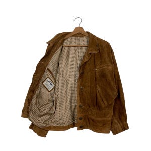 May include: A brown suede jacket with a button-up front and a collared neckline. The jacket has a pocket on the chest and a patterned interior lining. The jacket is hanging on a wooden hanger.
