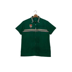 Puede incluir: Una camisa verde de manga corta con un diseño de rayas blancas y rojas en el pecho. La camisa presenta el logotipo de 7-Eleven en el pecho izquierdo y un cuello blanco.