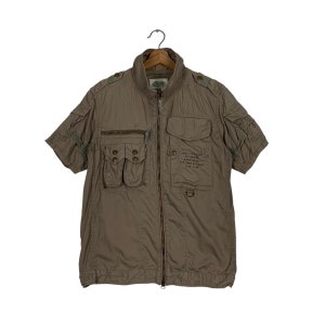 Puede incluir: Chaqueta verde oliva de manga corta con múltiples bolsillos y cremallera. La chaqueta tiene cuello alto y charreteras en los hombros. Texto impreso en el bolsillo delantero. Cuelga de una percha de madera.