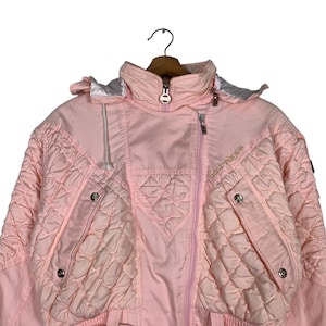 May include: A pink quilted ski jacket with a zip closure and the word "Ocunoboca" embroidered on the right side. The jacket has a hood and two pockets on the front.