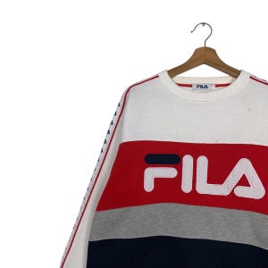 May include: A white, red, gray, and navy blue FILA sweatshirt. The sweatshirt has a color block design with the FILA logo in large white letters. The sleeves have a white stripe with small blue triangles.
