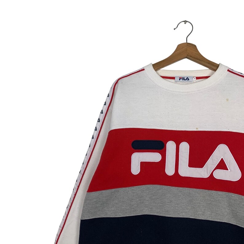 May include: A white, red, gray, and navy blue FILA sweatshirt. The sweatshirt has a color block design with the FILA logo in large white letters. The sleeves have a white stripe with small blue triangles.