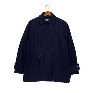 Könnte beinhalten: Marineblaue Jacke mit Kragen und langen Ärmeln. Die Jacke hat zwei Vordertaschen, einen Kordelzug in der Taille und geknöpfte Manschetten. Sie hängt an einem Holzbügel vor weißem Hintergrund.