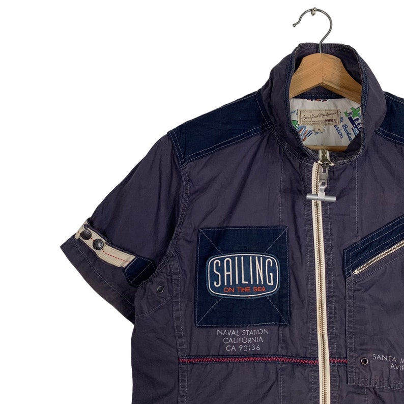 May include: A dark blue short-sleeved jacket with a zip fastening. The jacket has a patch on the front that says "Sailing on the Sea" and "Naval Station California CA 92136".