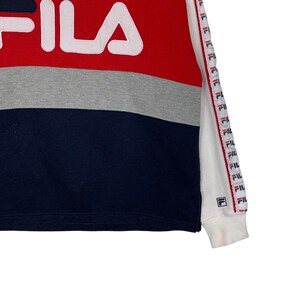 May include: A color-blocked sweatshirt in red, gray, and navy blue with the brand name "FILA" in white. The sleeve features a repeating logo pattern in red, white, and blue. The cuff is white.