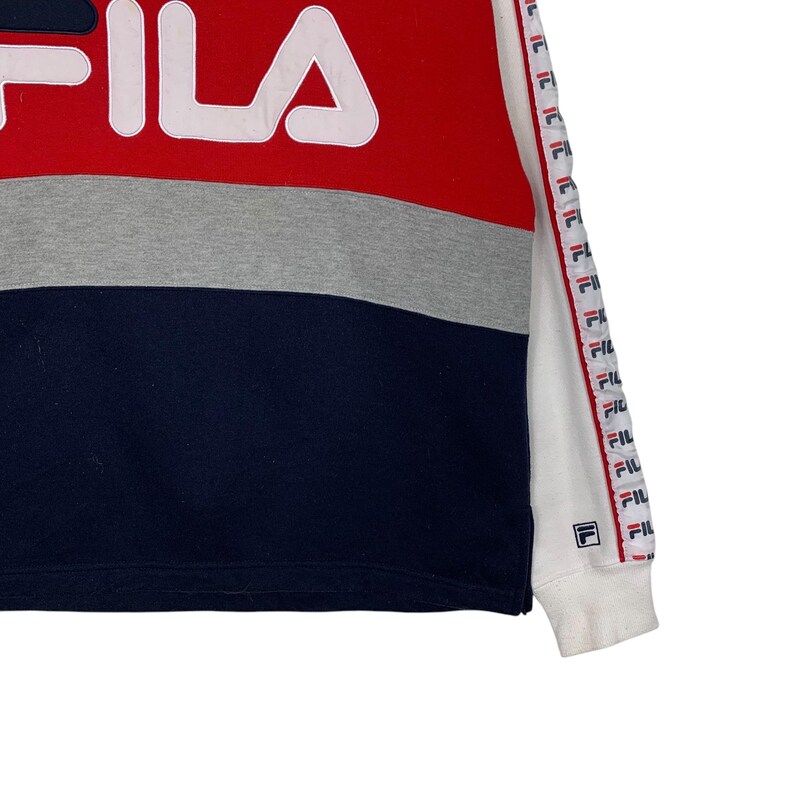 May include: A color-blocked sweatshirt in red, gray, and navy blue with the brand name "FILA" in white. The sleeve features a repeating logo pattern in red, white, and blue. The cuff is white.