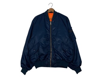 Puede incluir: Chaqueta bomber azul marino con forro naranja, colgada de una percha. La chaqueta tiene cierre de cremallera, dos bolsillos delanteros y un pequeño bolsillo en la manga izquierda. Los puños y la cintura son de color azul oscuro.