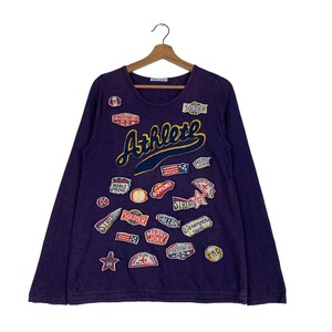 Op de afbeelding: Paarse longsleeve met verschillende kleurrijke patches en het woord "Athlete" in een groot, geel en blauw lettertype.