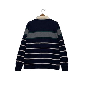 May include: Navy blue long-sleeve rugby shirt with white horizontal stripes. The shirt features a cream-colored collar and gray and green horizontal stripes across the chest. The shirt is hanging on a wooden hanger.
