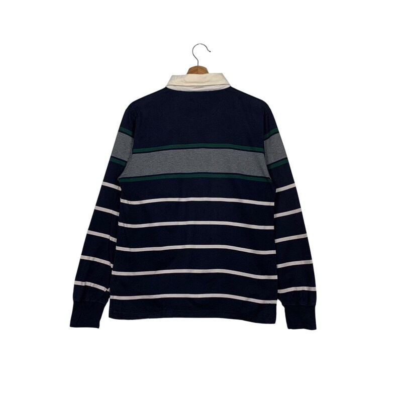 May include: Navy blue long-sleeve rugby shirt with white horizontal stripes. The shirt features a cream-colored collar and gray and green horizontal stripes across the chest. The shirt is hanging on a wooden hanger.