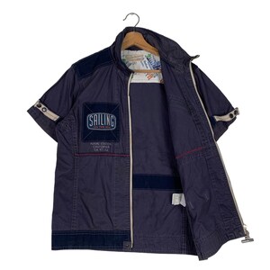 May include: A navy blue short-sleeved jacket with a zip fastening. The jacket has a patch on the left chest that says "Sailing on the Bay" and "Naval Station California CA 92138".
