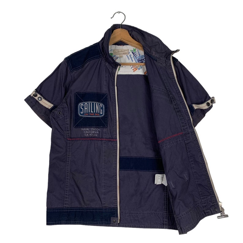 May include: A navy blue short-sleeved jacket with a zip fastening. The jacket has a patch on the left chest that says "Sailing on the Bay" and "Naval Station California CA 92138".